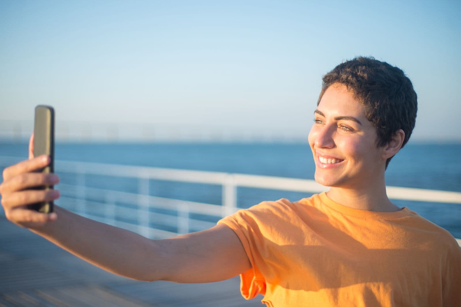 Smiling traveller in an orange t-shirt holding up her phone by a sunlit blue sea